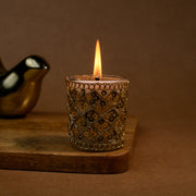 Lit decorative gold-patterned candle in a glass holder on wooden tray, brown background